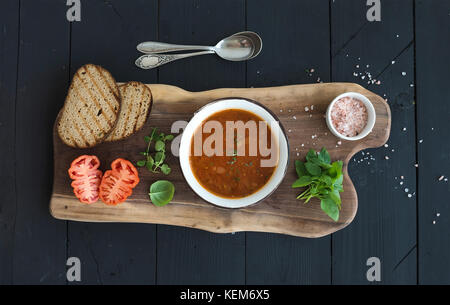 Roasted tomato soup with fresh basil, spices and bread in vintage metal bowl on wooden board over black background Stock Photo
