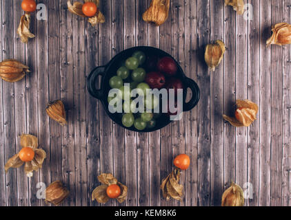 Fresh juicy group of organic physalis, green grapes and gooseberries in black bowl on wooden table. Flat lay style. Stock Photo