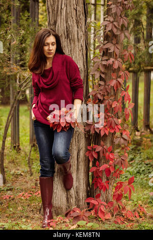 Young woman (brunette) in jeans, a sweater and crimson rubber boots is walking in an autumn park. Selective focus. Stock Photo