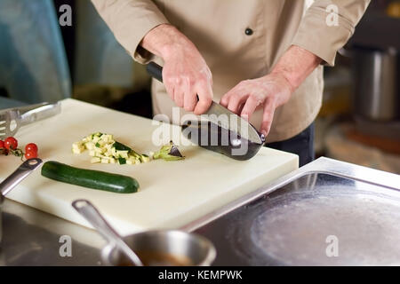 Chef cutting eggplant by steel knife in restaurant. Male chef cutting eggplant on cutting board next to sliced zucchini. European cuisine concept. Stock Photo