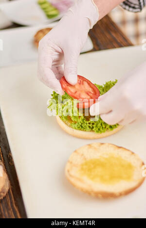 Chef adding tomatoes on burger. Cook preparing hamburger at professional kitchen. Cooking and cuisine concept. Stock Photo