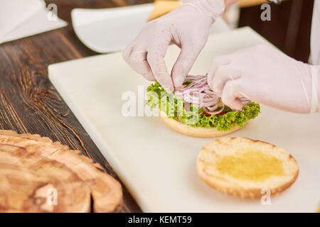Chef hands adding onion on burger. The process of cooking fresh delicious hamburger at kitchen by chef. Chef working at kitchen. Stock Photo
