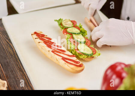 Chef hands cutting sausage to hot dog. The process of cooking long sandwich at kitchen. Chef at work, restaurant. Stock Photo