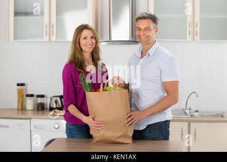 Portrait Of Happy Couple Holding Grocery Bag With Vegetables In Kitchen Stock Photo