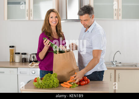 Happy Mature Couple Removing Vegetables From Grocery Bag In Kitchen Stock Photo