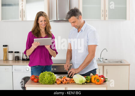Wife Helping Her Husband For Preparing Food With Digital Tablet Stock Photo