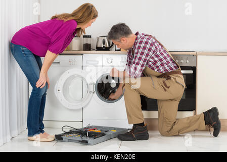 Woman Looking At Repairman Checking Washing Machine In Kitchen Stock Photo