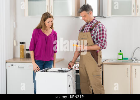 Woman Looking At Male Technician Checking Washing Machine In Kitchen Stock Photo