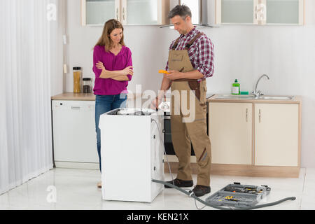 Woman Looking At Male Technician Checking Washing Machine In Kitchen Stock Photo
