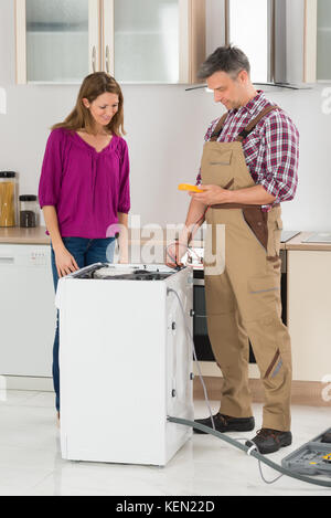 Woman Looking At Male Technician Checking Washing Machine In Kitchen Stock Photo