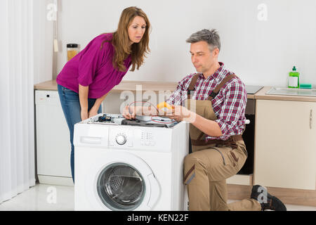 Woman Looking At Male Technician Checking Washing Machine In Kitchen Stock Photo