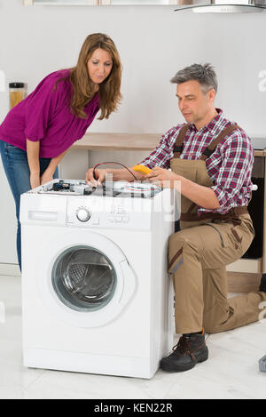 Woman Looking At Male Technician Checking Washing Machine In Kitchen Stock Photo