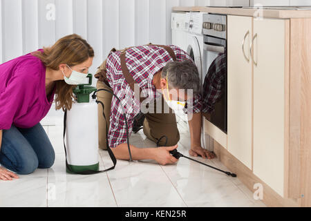 Mature Woman Looking At Male Worker Spraying Insecticide In Kitchen Stock Photo
