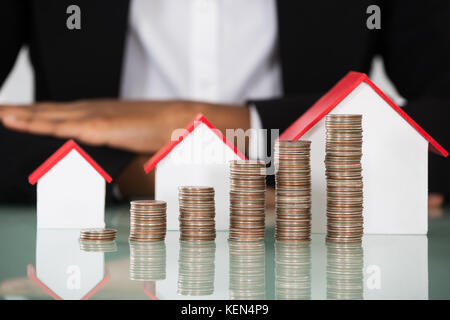 Close-up Of Businesswoman With Different Size Of House Model And Stacked Coins On Desk