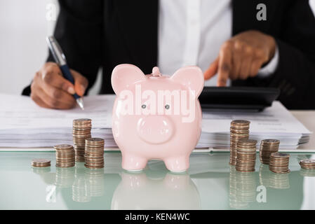 Close-up Of Businesswoman Calculating Tax With Piggybank And Stack Of Coins On Office Desk Stock Photo