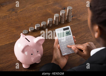 Close-up Of A Businesswoman Using Calculator With Piggybank And Stack Of Coins On Desk Stock Photo