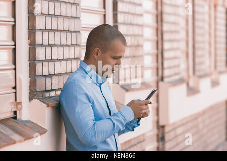 Portrait of a smiling handsome man in sweater using a mobile phone while leaning on a wall outdoors. Texting message Stock Photo