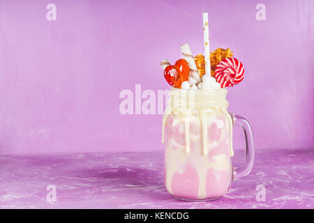 Freakshake from pink smoothie, cream. Monstershake with lollipops, waffles and marshmallow. Extreme milkshake in a Mason jar. Pink purple background. Stock Photo