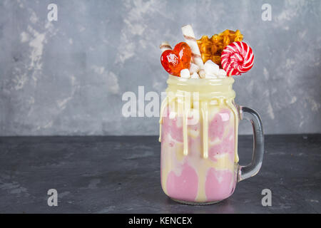 Freakshake from pink smoothie, cream. Monstershake with lollipops, waffles and marshmallow. Extreme milkshake in a Mason jar. Gray dark concrete backg Stock Photo