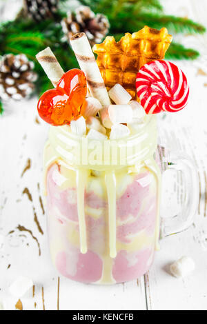 Freakshake from pink smoothie, cream. Monstershake with lollipops, waffles and marshmallow. Extreme milkshake in a Mason jar. White wooden table. Atmo Stock Photo