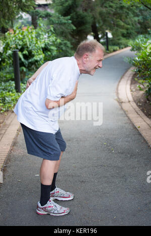 Closeup portrait, old man clutching chest, having heart pain, after strenuous workout, isolated outdoors, green trees background. Myocardial infarctio Stock Photo