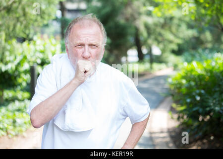 Closeup portrait, senior guy holding towel, very tired, exhausted from over exertion, coughing catching breath, isolated outdoors outside green trees  Stock Photo