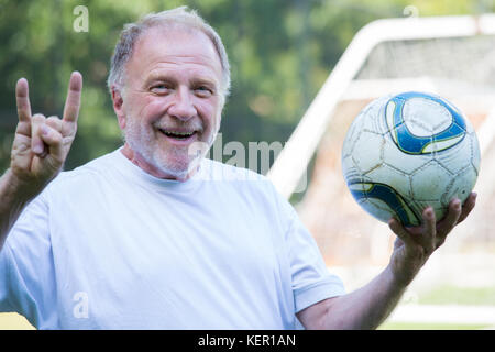 Closeup portrait, elderly man holding up soccer ball and showing off horns sign with hands, isolated soccer goal post outdoors background Stock Photo