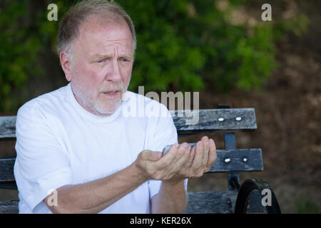 Closeup portrait of bold elderly man in white shirt, squinting, checking smartphone, sending text message, seated on a bench, isolated outdoor backgro Stock Photo