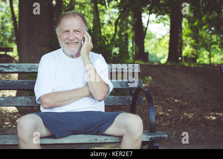 Closeup portrait, older, happy ecstatic man sitting on bench talking on cell phone, isolated outdoors background Stock Photo