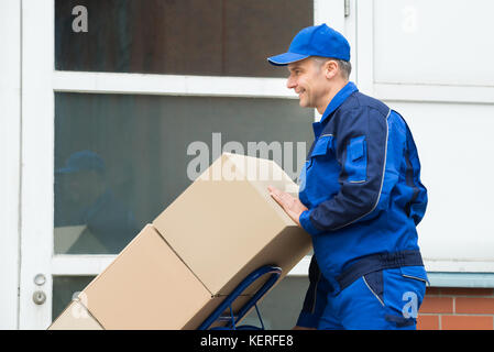 Mature Happy Delivery Man Carrying Boxes On A Hand Truck On Street Stock Photo