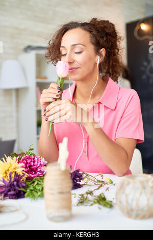 Portrait of pretty-mixed race woman joyfully smelling flowers arranging beautiful bouquet  at desk in modern apartment and listening to music Stock Photo