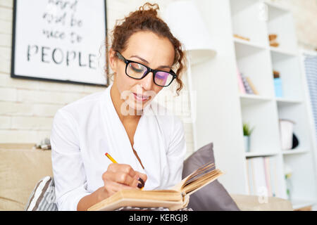 Portrait of elegant young woman wearing glasses making notes in notebook while sitting in comfortable armchair Stock Photo