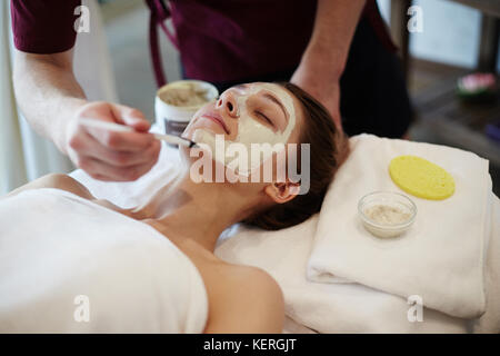 Closeup of unrecognizable male cosmetologist applying face mask with brush to face of beautiful woman lying on massage table in SPA center Stock Photo