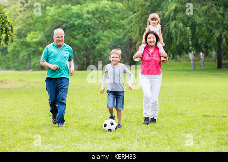 Happy Family Playing Soccer Game With Grandchildren Together In Park. Running For Ball Stock Photo