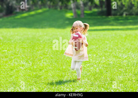 Little Happy Girl Holding Ball While Playing American Football In The Park Stock Photo