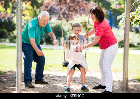 Family Having Fun With Kids Having Ride On Swing In The Park Stock Photo