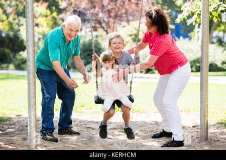 Senior Grandparents Having Fun With Kids On Swing In The Park Stock Photo