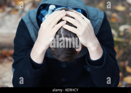 Young man covers his face with his hands in grief or pain. He is sitting outdoor. Stock Photo