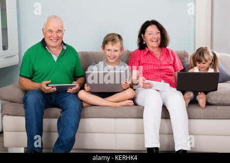 Family Sitting On Couch With Their Laptop Computers And Tablets At Home Stock Photo