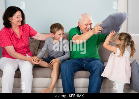 Older Happy Grandparents Having Pillow Fight With Kids On Couch Together At Home Stock Photo