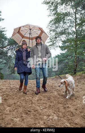 Young trendy couple, male & female wearing winter clothes and sheltering under an umbrella walking with their dog on a sandy path in a woodland area Stock Photo