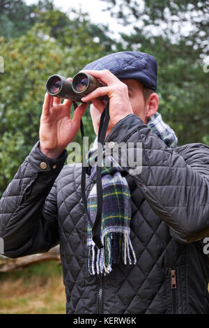 Young male dressed in winter clothing looking through binoculars in a woodland setting Stock Photo