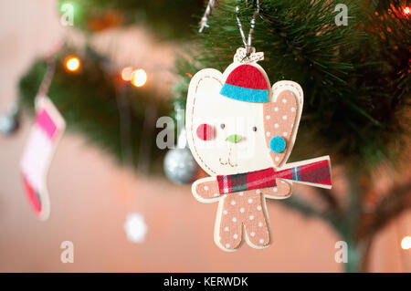 A wooden Christmas toy in the form of a cartoon cat dressed in winter hat, red checkered scarf and baby romper with polka dots print is hanging on a b Stock Photo