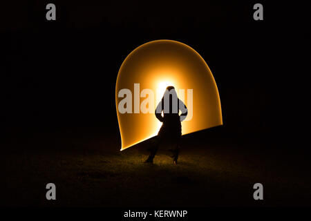 Silhouette figure of lone woman in coat standing isolated in the dark, black background with unexplained, golden light waves in arc. Light painting UK. Stock Photo