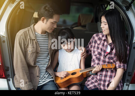 happy little girl playing ukulele with asian family sitting in the car for enjoying road trip and summer vacation Stock Photo
