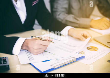 Asian businesswomen holding a pen and analysis documents on office table with laptop computer and graph financial diagram working in the background Stock Photo