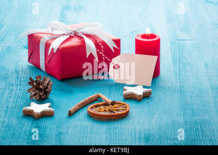 Christmas scenery with a red gift box, a lit candle, gingerbread cookies and cinnamon sticks on a blue wooden table Stock Photo