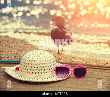 Beach scene. Sun straw hat and sunglasses lying on the sea  on the beach. Seagull walking on the beach back to camera Stock Photo