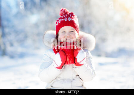 Child drinking hot chocolate with marshmallows in snowy winter park. Kid with cup of warm cocoa drink on Christmas vacation. Little girl playing in sn Stock Photo