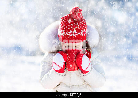 Child drinking hot chocolate with marshmallows in snowy winter park. Kid with cup of warm cocoa drink on Christmas vacation. Little girl playing in sn Stock Photo